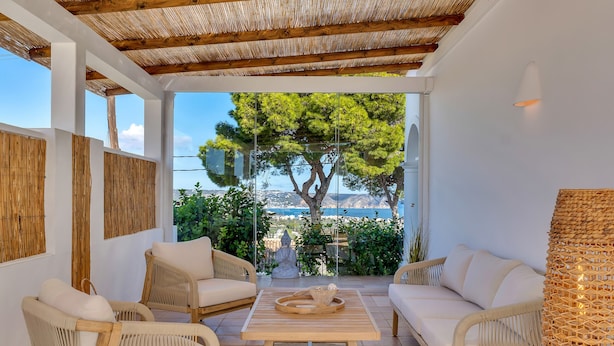 A sunlit patio with wicker furniture, a wooden table, and a view of a tree and landscape through a glass door. - Casa Iris (Javea / Xàbia)