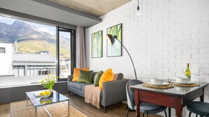 Una sala de estar moderna con sofá, mesa de centro y mesa de comedor. Hay dos cuadros en la pared, una lámpara y vistas de una montaña a través de un ventanal grande.