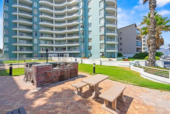 Un espace patio avec un barbecue en briques et des tables de pique-nique, entouré d’une clôture blanche et d’un immeuble imposant doté de nombreux balcons.