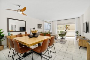 Un espace repas moderne avec une table en bois et des chaises marron, un ventilateur de plafond et une grande fenêtre avec des stores.