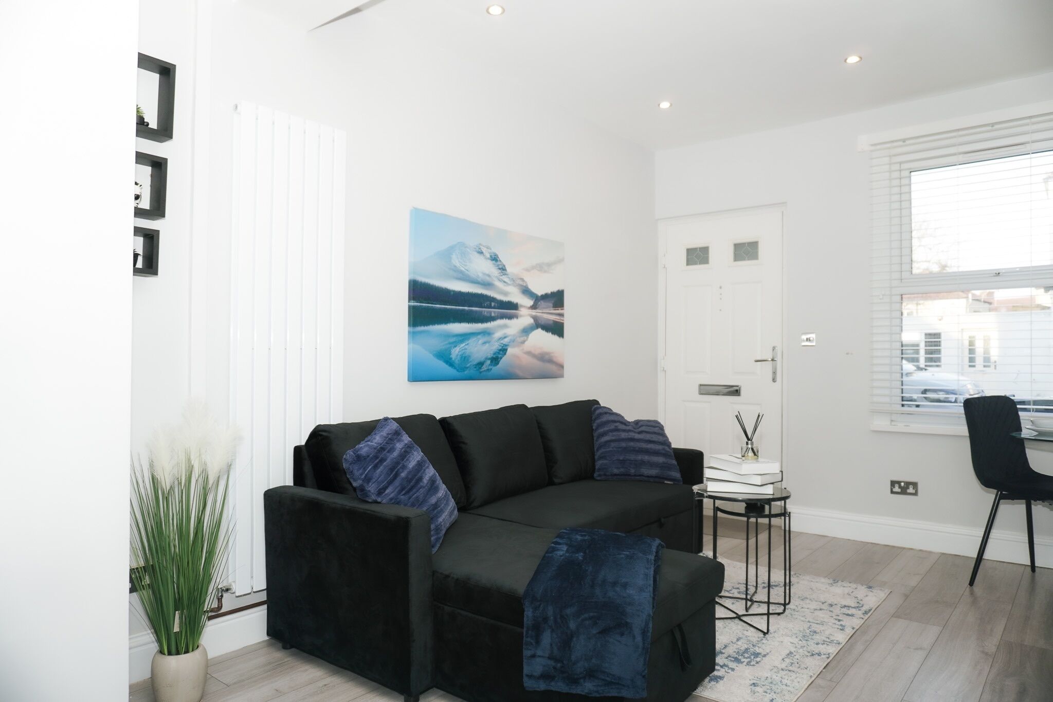 A modern living room with a black sofa, a painting of a mountain landscape, a potted plant, and a window with blinds.
