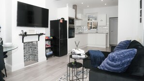 A modern living room with a black sofa, a flat-screen TV mounted on the wall, a kitchen area with white cabinets, and a dining table with a patterned rug. - 3 Bed-sleeps 7-garden-parking-town Centre (Dartford)