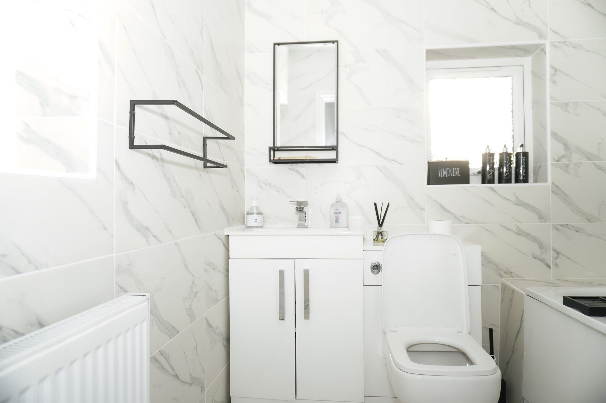 A modern bathroom with white marble tiles, a toilet, a sink with a mirror, and a shelf with toiletries.