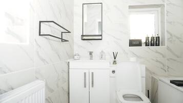 A modern bathroom with white marble tiles, a toilet, a sink with a mirror, and a shelf with toiletries.