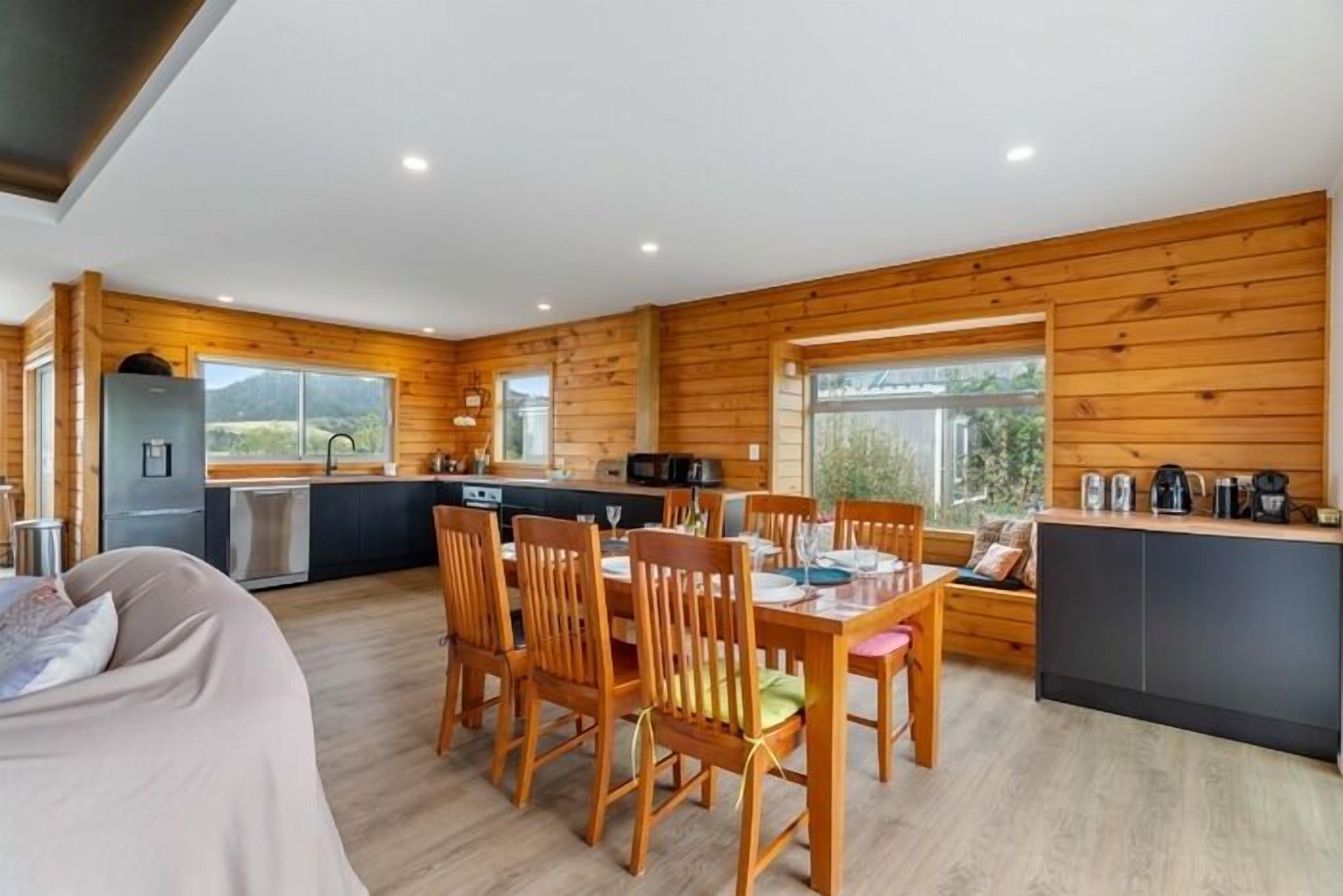 A modern kitchen with wooden walls, a dining area with wooden chairs, and a large window offering a view of the outdoors.
