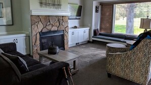 A living room with a stone fireplace, a television, a sofa, a patterned armchair, and a window with blinds.