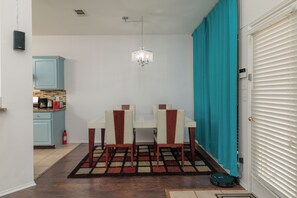Un coin repas avec une table et des chaises, un lustre, une cuisine en arrière-plan et un rideau de couleur turquoise.