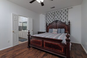 Une chambre à coucher avec un lit en bois, un ventilateur de plafond et une salle de bain visible à travers une porte ouverte.