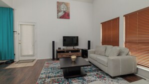 En stue med sofa, sofabord, fjernsyn, højtalere og en dør med cremefarvede gardiner.