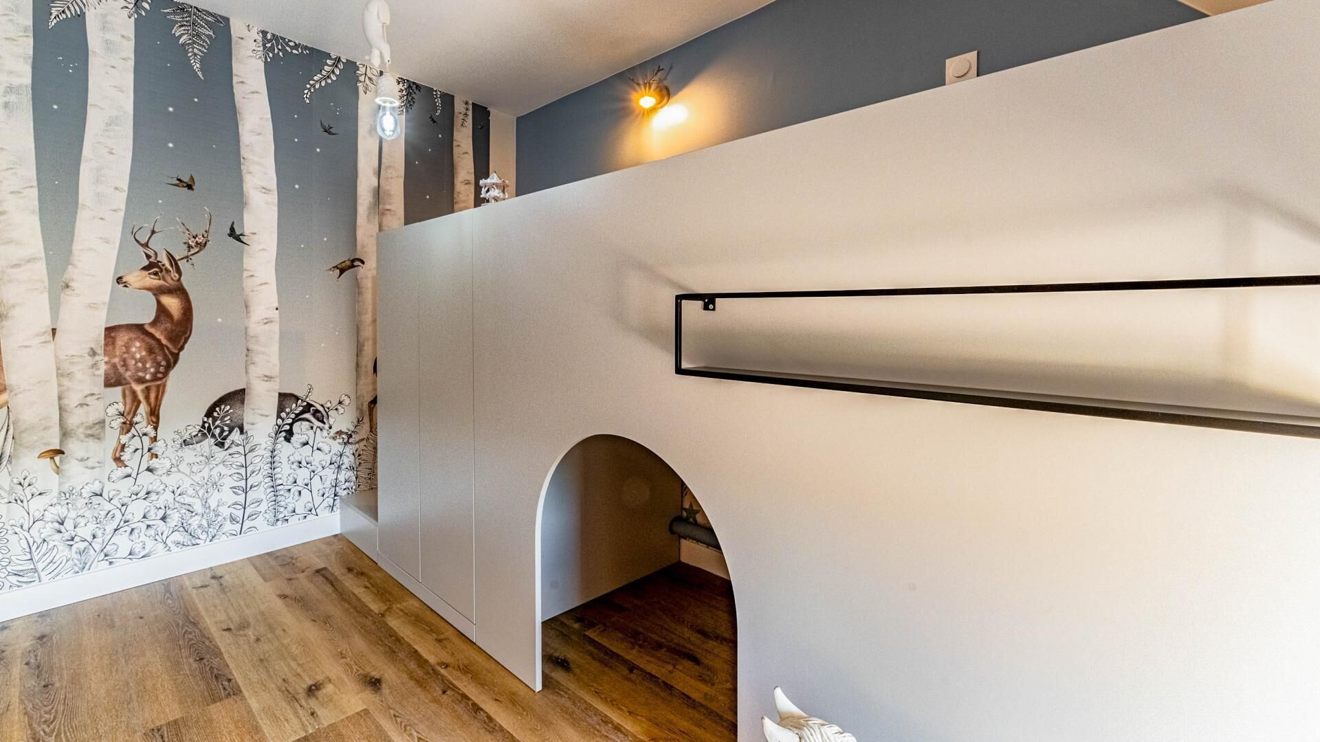 A room with a wooden floor, a white wall with a black shelf, and a mural of deer and leaves on the wall.