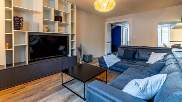 A modern living room with a large flat-screen TV, a dark sofa, and a wooden floor.
