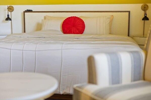 Una cama bien hecha con un cojín decorativo rojo, un cabecero blanco y dos lámparas de noche.