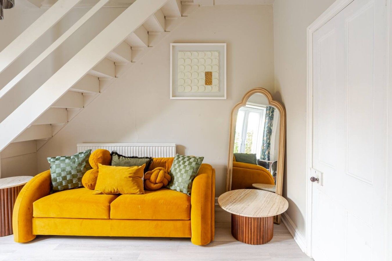 A room with a yellow sofa, a mirror, a framed artwork, and a staircase.