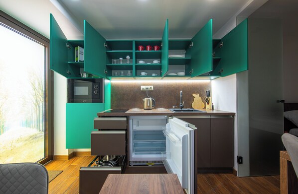 Una cocina compacta con microondas integrado, refrigerador y lavamanos. Hay armarios abiertos con vajilla y una tetera sobre la encimera.