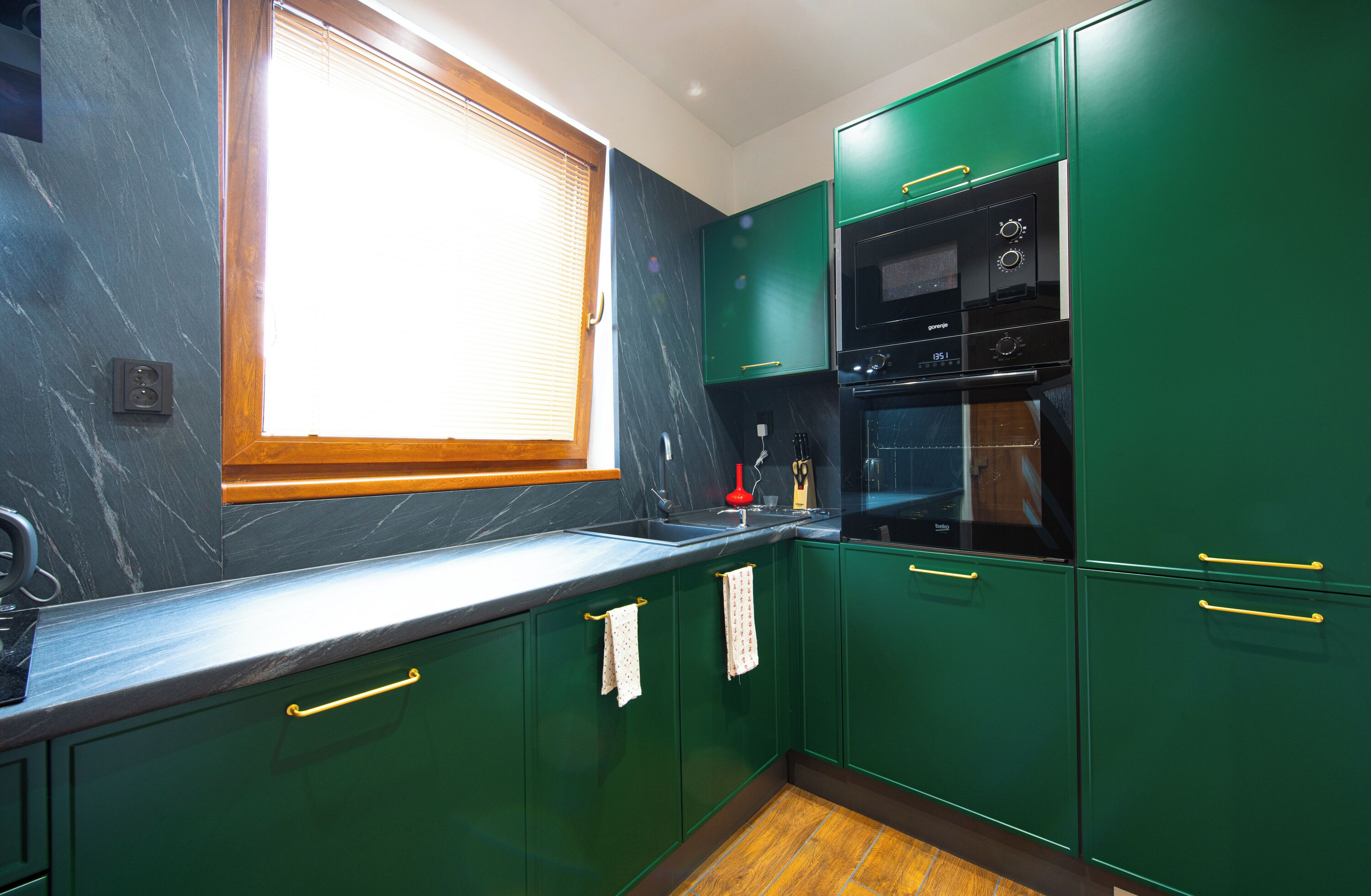 A kitchen with green cabinets, a black countertop, and a window with blinds.