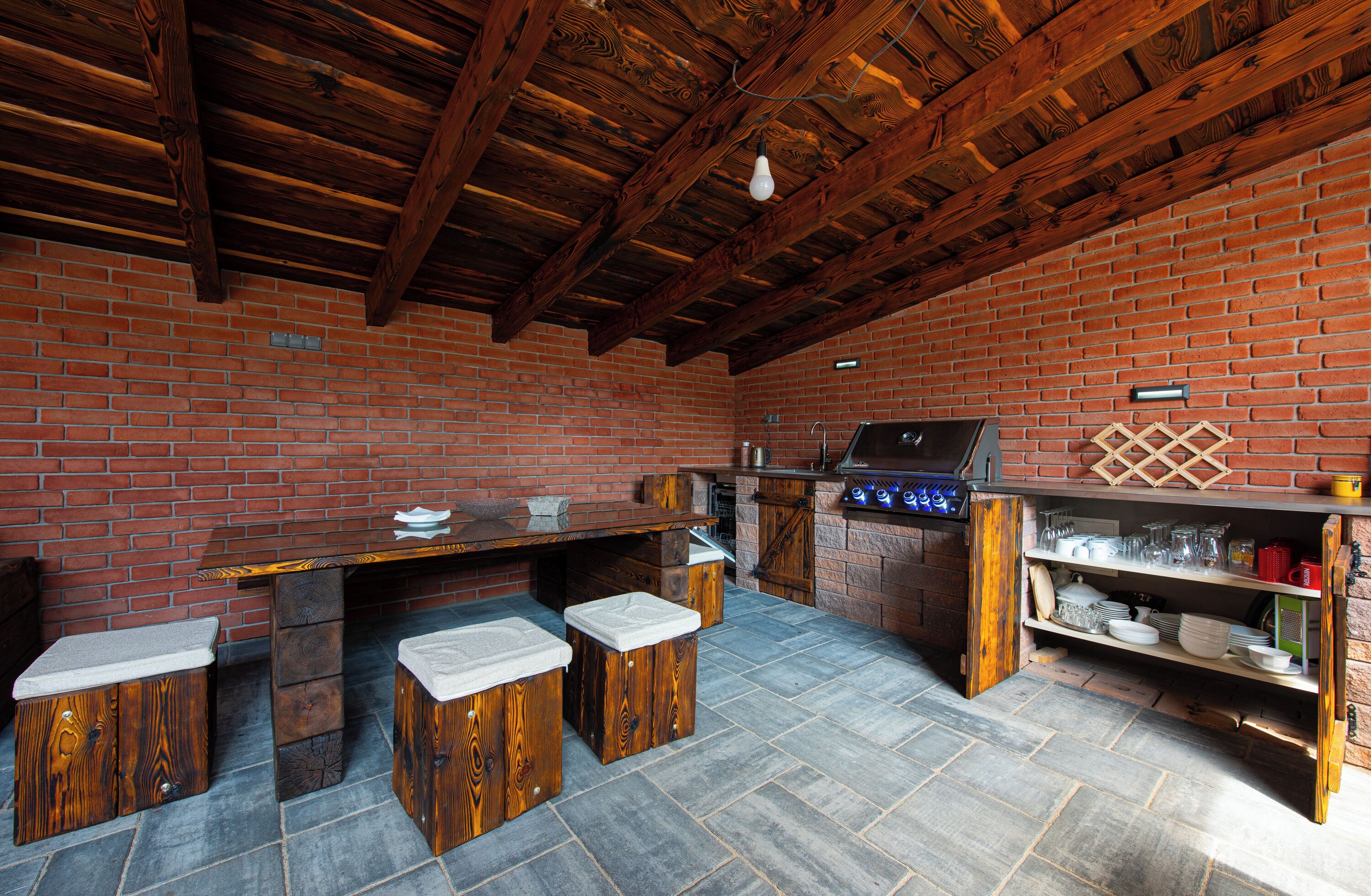 A rustic outdoor kitchen with a brick wall, wooden beams, and a stone floor.