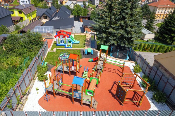 Ein Spielplatz mit verschiedenen Spielgeräten, umgeben von einem Zaun und Wohnhäusern im Hintergrund.