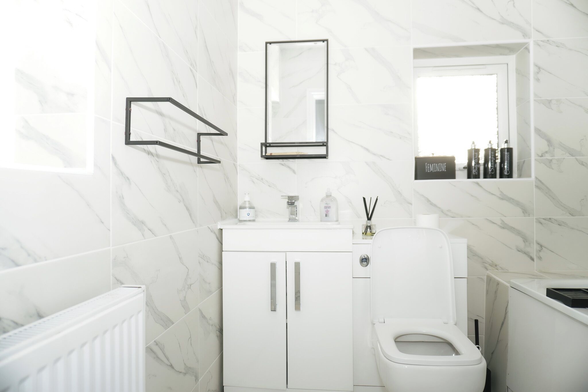 A modern bathroom with a white marble wall, a toilet, a sink with a mirror, and a shelf with toiletries.