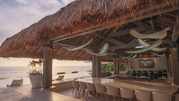 A beachside bar with a thatched roof, a long counter with bar stools, and a view of the ocean.
