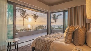 A bedroom with a large window offering a view of the ocean and palm trees at sunset.