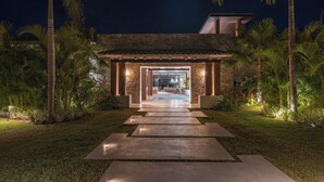 A modern house with a stone facade, a pool, and a well-lit entrance surrounded by palm trees.