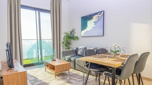 A modern living room with a sofa, a wooden coffee table, a dining table with chairs, a TV on a wooden stand, and a large window with curtains. - Bloomfields 1Br Binghatti Heights (Dubai)