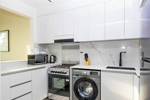 A compact kitchen with white cabinets, a microwave, a kettle, a stove, and a washing machine. - Bloomfields 1Br Binghatti Heights (Dubai)
