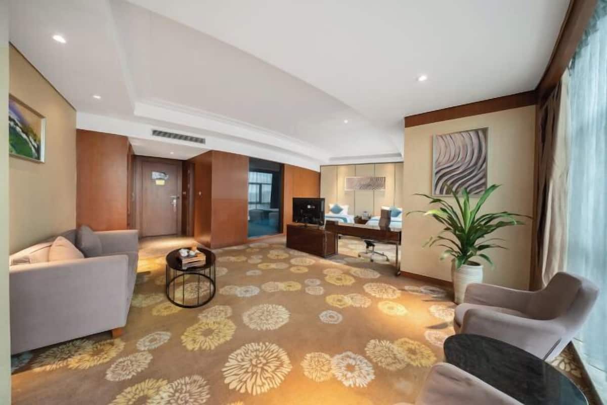 Habitación de hotel moderna con sofá, sillones, escritorio, televisor y un amplio ventanal.