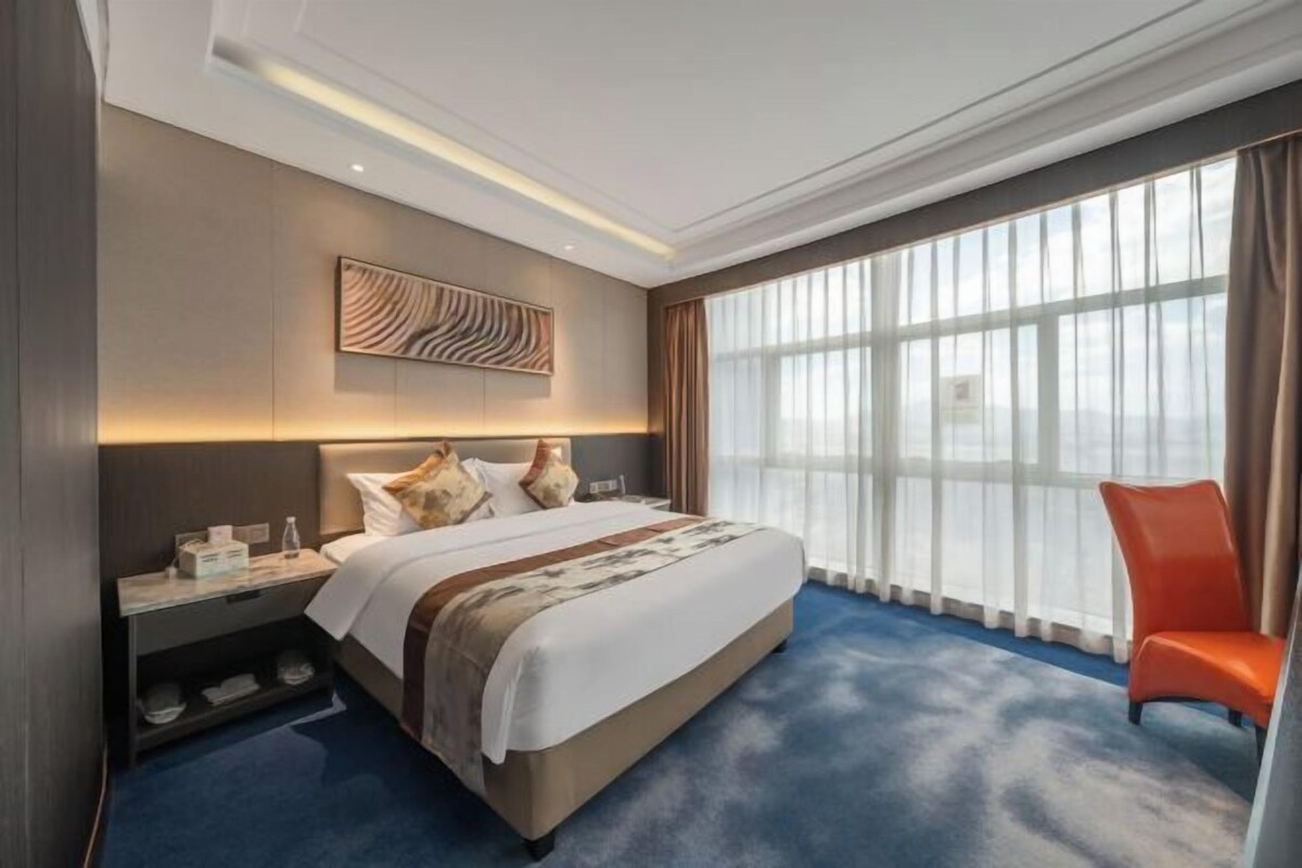 A hotel room with a large bed, a nightstand, a chair, a large window, and a wall art piece.