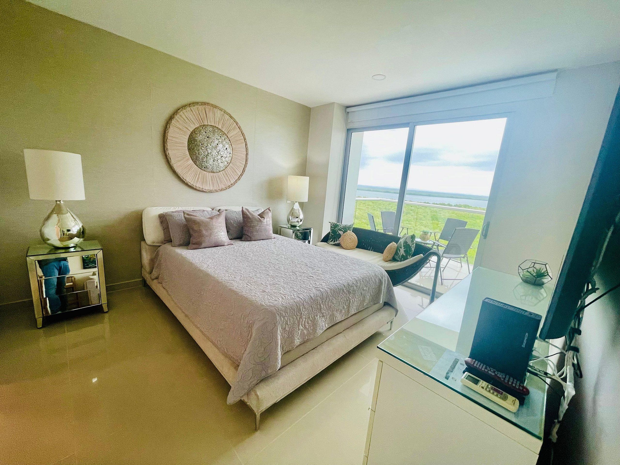 Un dormitorio con cama, dos mesitas de noche, una silla, una mesa de vidrio con una computadora portátil y un ventanal con vistas.