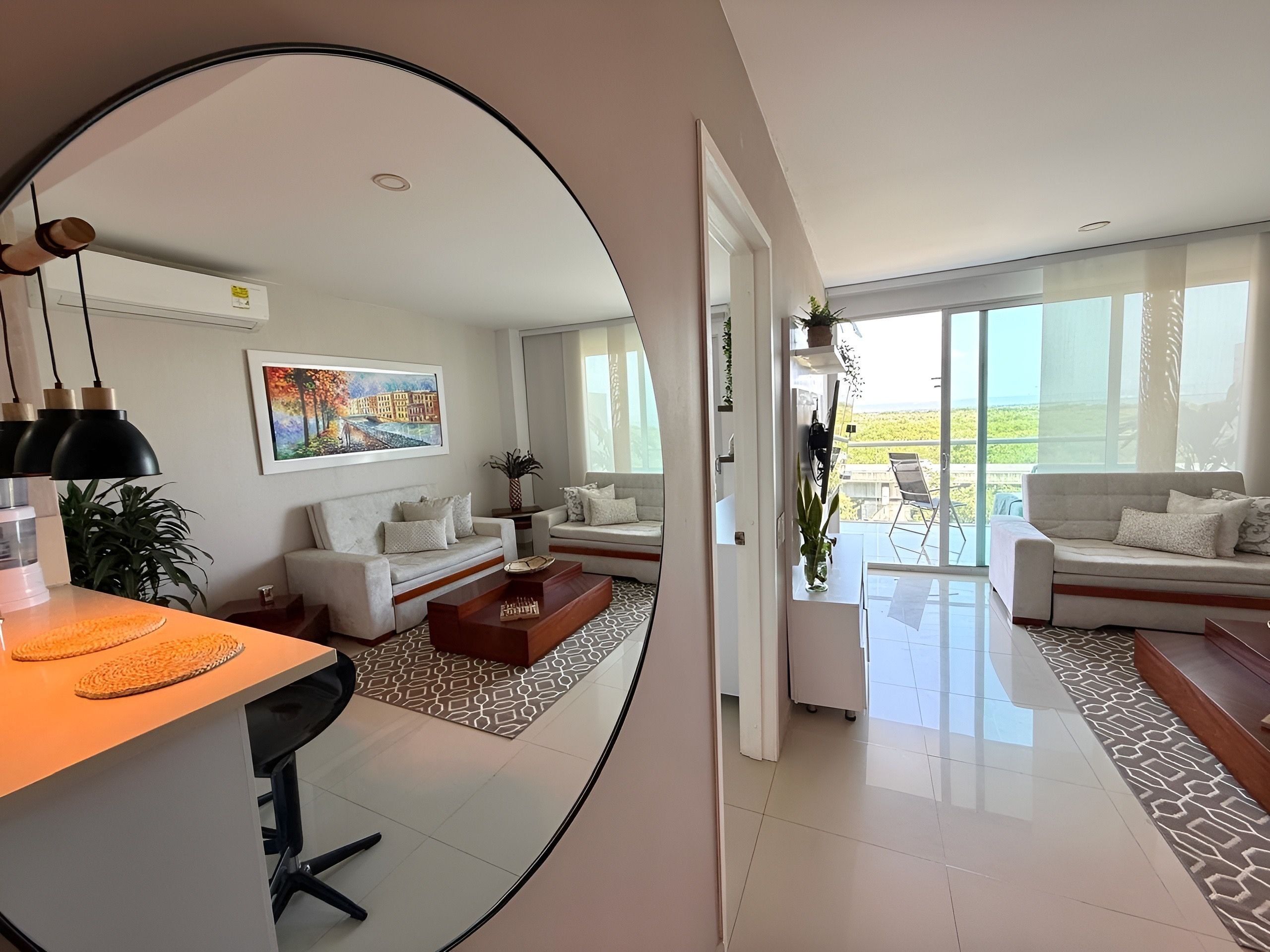 A modern living room with a sofa set, a coffee table, and a dining area with a table and chairs. There is a large window providing a view of the outdoors.