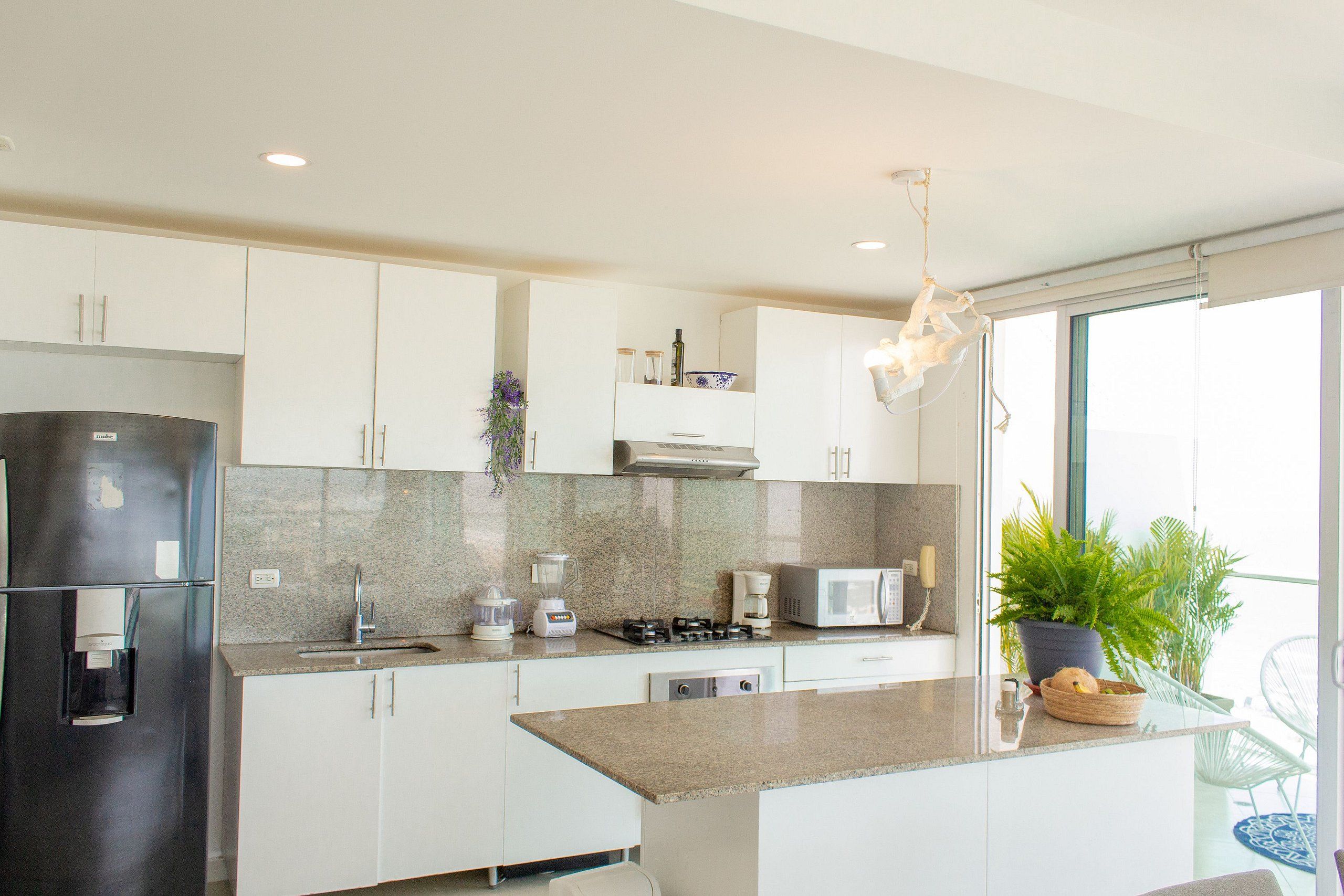 A modern kitchen with white cabinets, a granite countertop, a stainless steel refrigerator, a microwave, a stove, and a sink. There is a potted plant on the countertop and a hanging light fixture above the island.