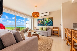 A spacious living room with a large window, a grey sofa, a coffee table, and a dining area with wooden chairs. - Condominio frente al mar  Morros Epic. (Cartagena)