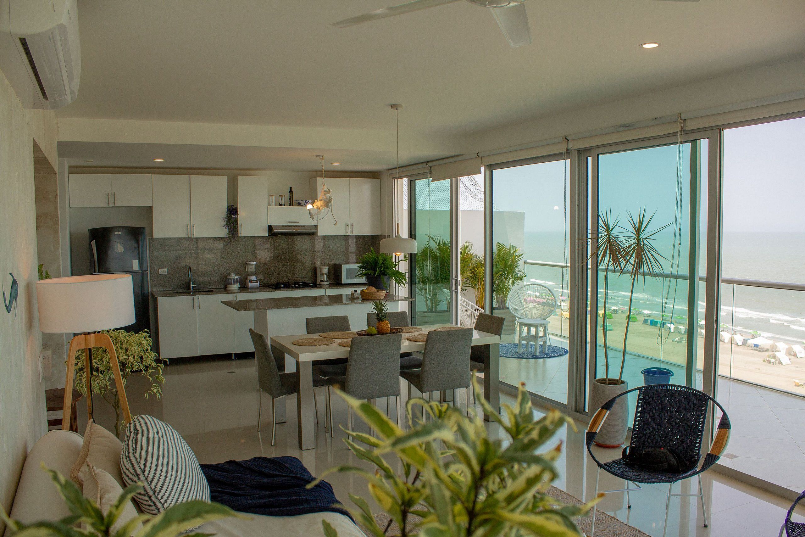 Una cocina moderna con armarios blancos, un comedor con mesa y sillas, y ventanas amplias con vista a la playa.