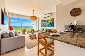 A modern living room with a large window, a dining area with a kitchen counter, and a view of the outdoors.