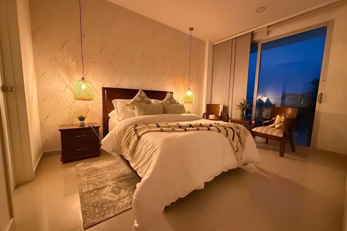 Une chambre à coucher avec un grand lit, des tables de chevet, une commode, une chaise et une vue sur l’extérieur.