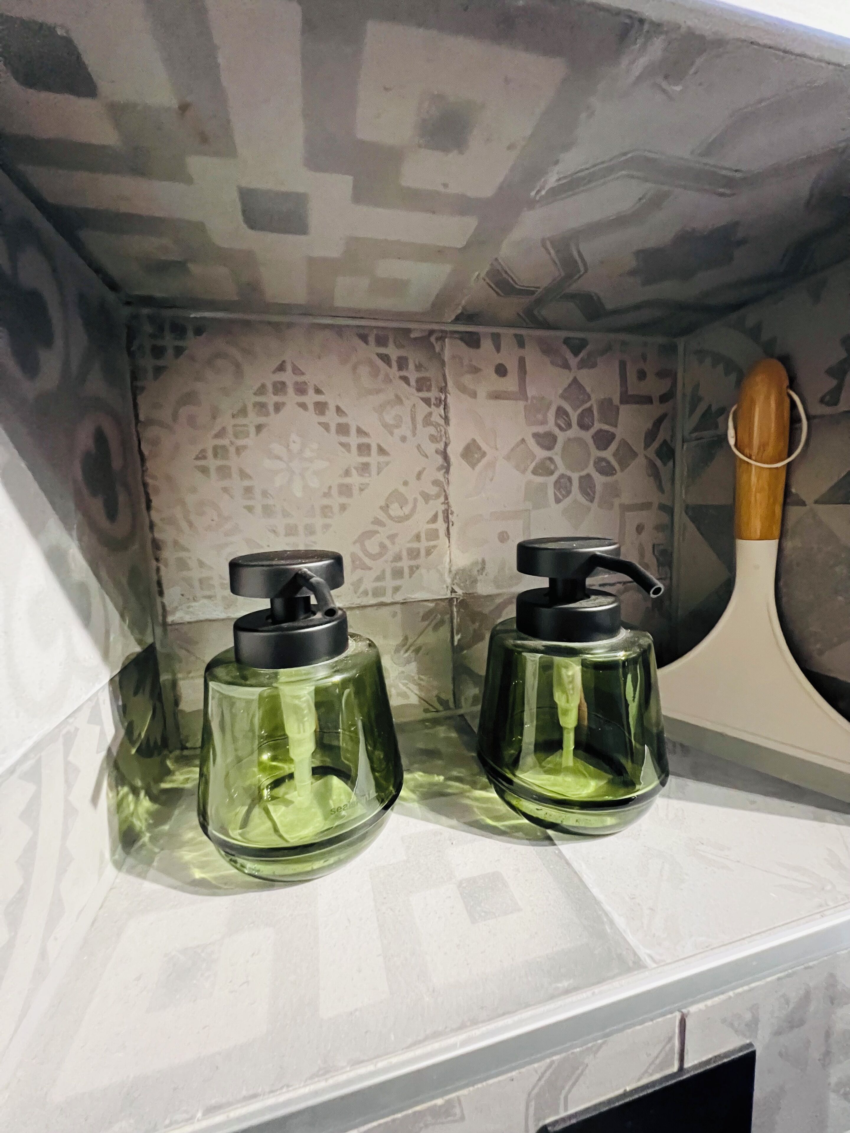 Une salle de bain avec un mur recouvert de carreaux décoratifs et deux bouteilles en verre vert dotées de pompes noires, posées sur un comptoir blanc.
