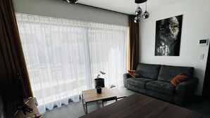 A modern living room with a dark wooden coffee table, a grey sofa with a brown cushion, a black floor lamp, and a large window with sheer curtains. - Liège city charm - Opera view (Liège)