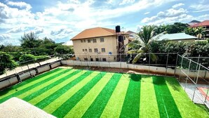 A rooftop with a green artificial turf field, surrounded by a metal fence, with buildings and trees in the background.