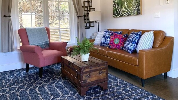 Un salon avec un fauteuil rose, un canapé marron, une table basse en bois ornée d’une plante et un tapis à motifs.