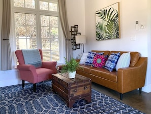 Un salon avec un fauteuil rose, un canapé marron, une table basse en bois ornée d’une plante et un tapis à motifs.
