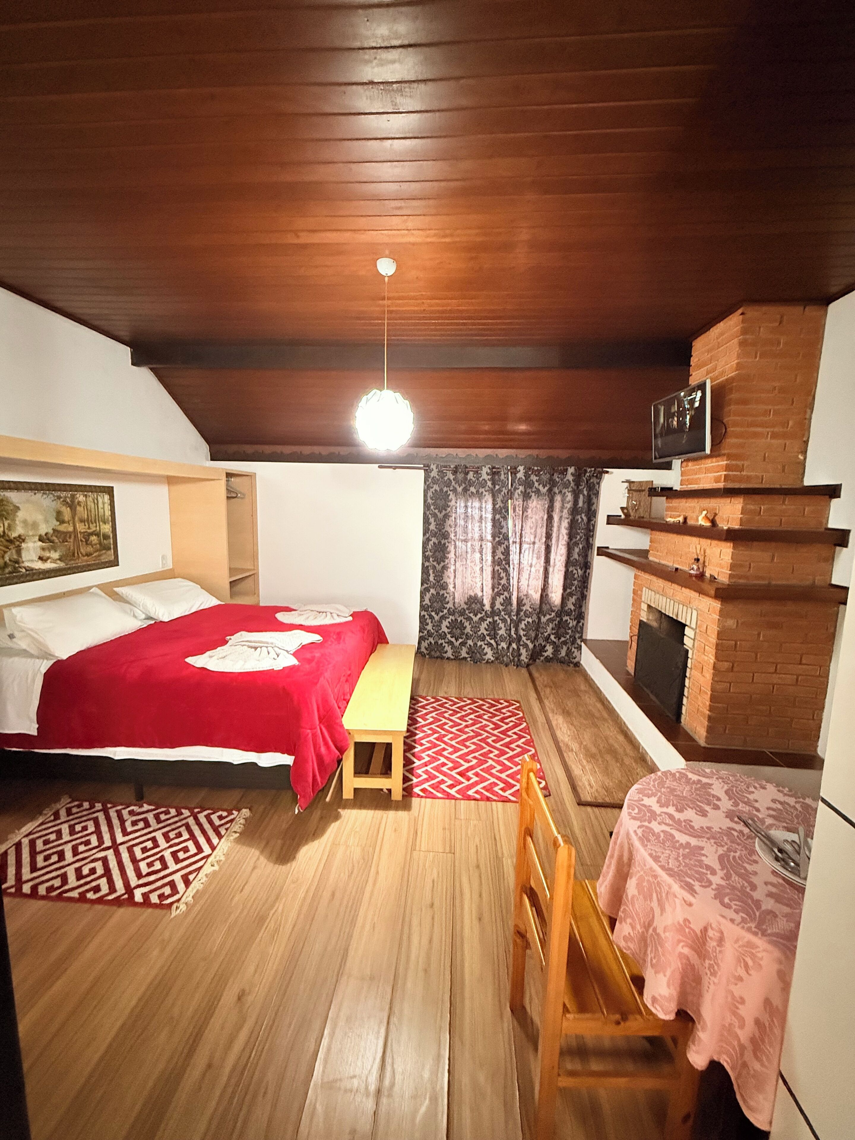 Una habitación acogedora con techo de madera, una cama con manta roja, una mesita, un banco, una chimenea y un televisor montado en la pared.