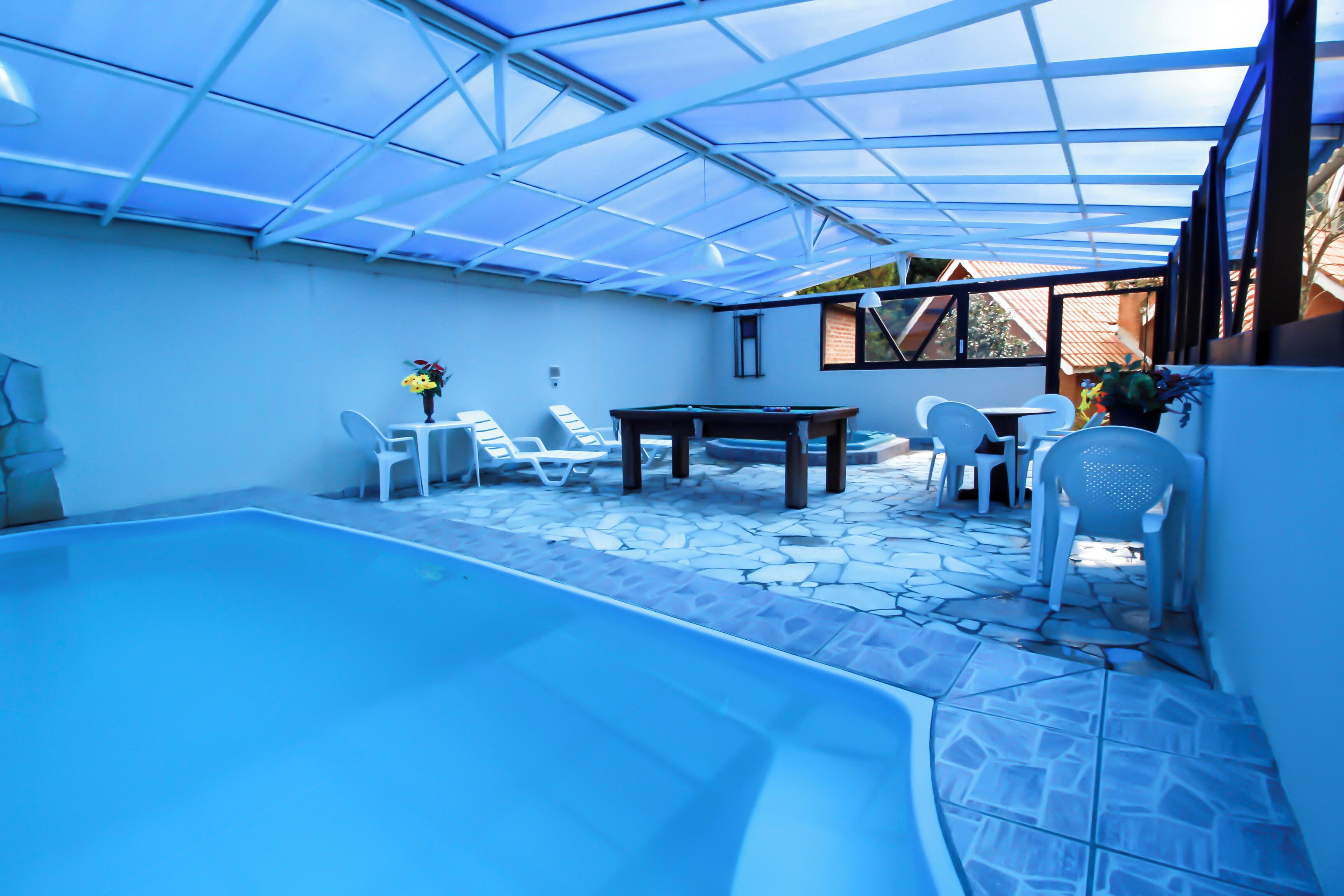 A covered outdoor swimming pool area with a table and chairs, stone flooring, and a glass ceiling.