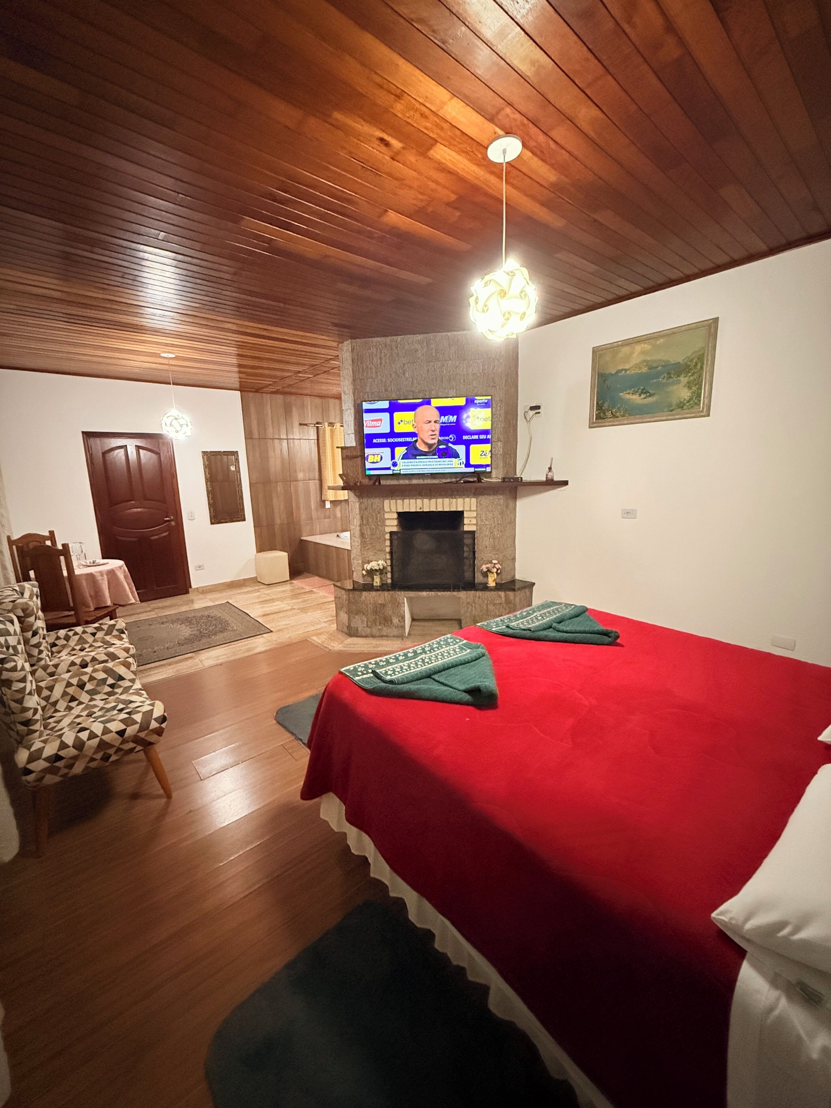 A room with a fireplace, a television mounted above it, a bed with a red bedspread, and a patterned armchair.