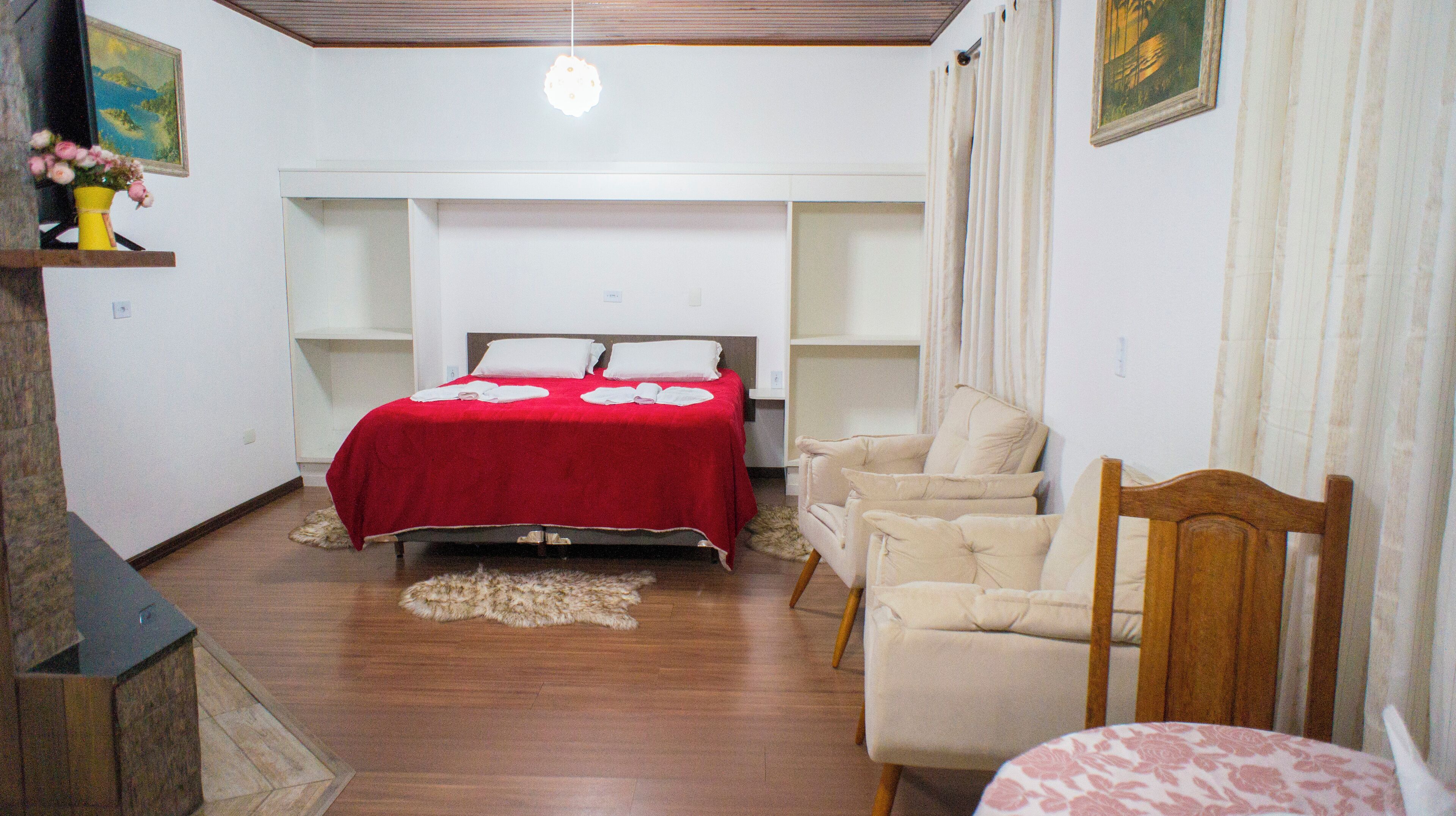 A bedroom with a bed, a chair, a table, and a small shelf with a vase of flowers.