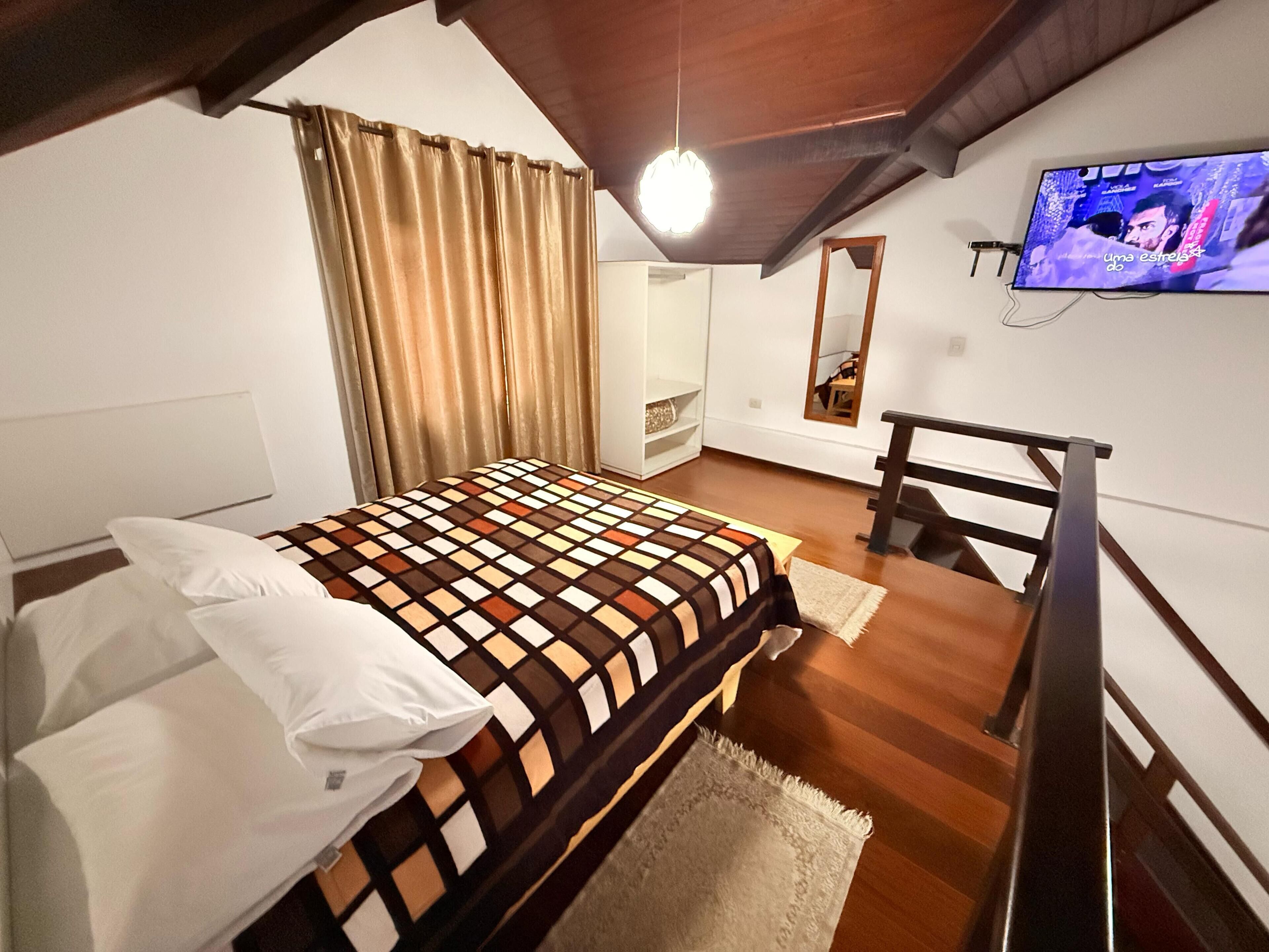 A bedroom with a bed, a television, a chair, and a wooden ceiling.