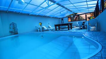 A swimming pool with a glass ceiling, white lounge chairs, and a table with chairs.