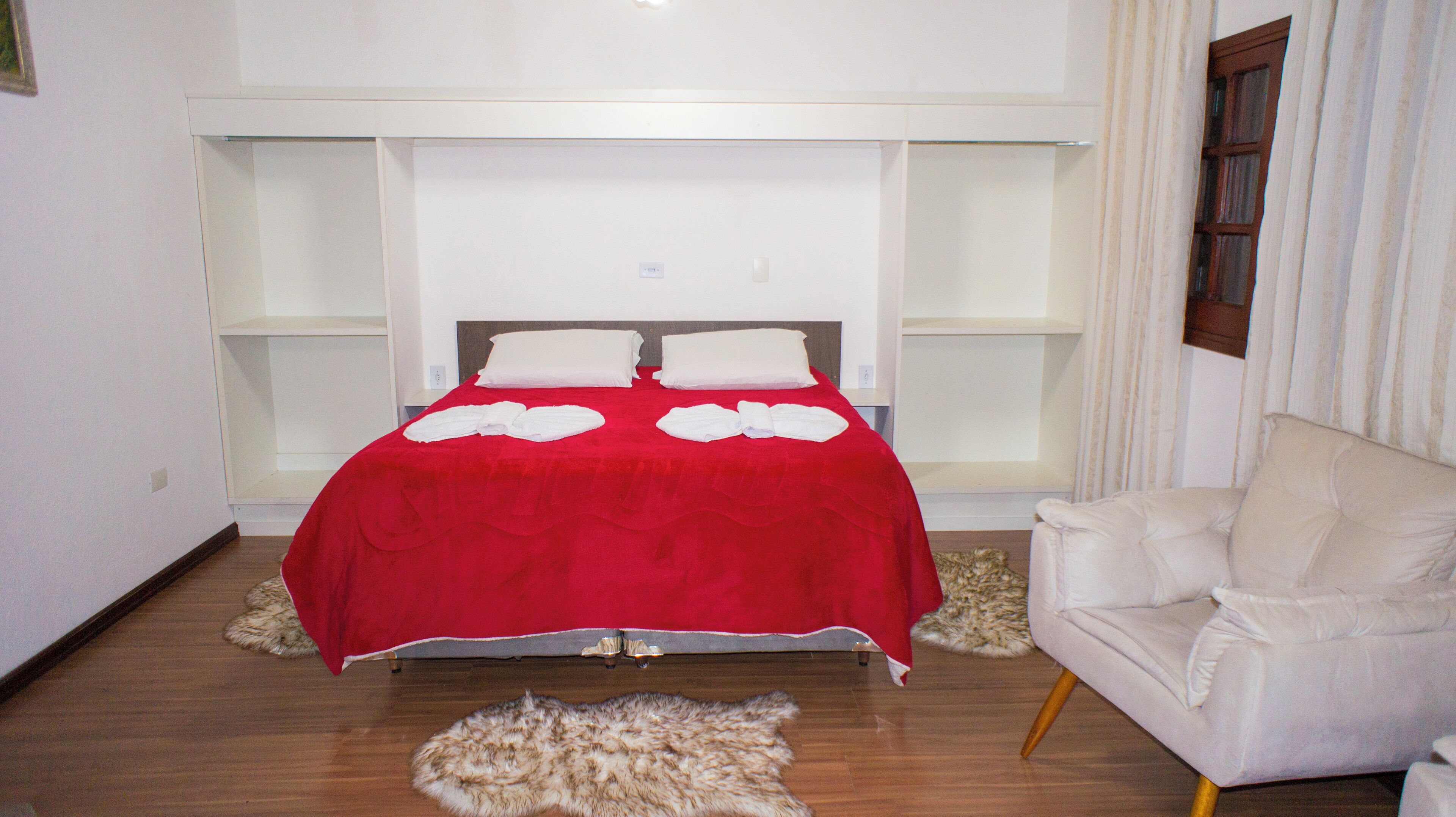 A bedroom with a red bedspread, white pillows, a white chair, and a fur rug.