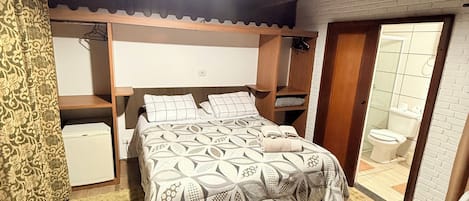 A bedroom with a bed, a bedside table, a hanging light fixture, a wardrobe, and a bathroom visible through an open door.