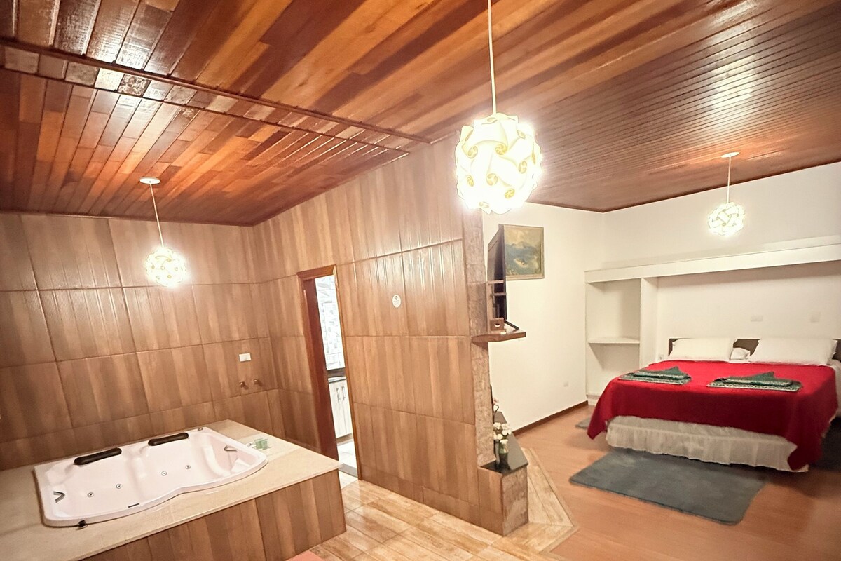 A room with a wooden ceiling, a large bathtub, a bed with a red blanket, and a small table with a pink tablecloth.